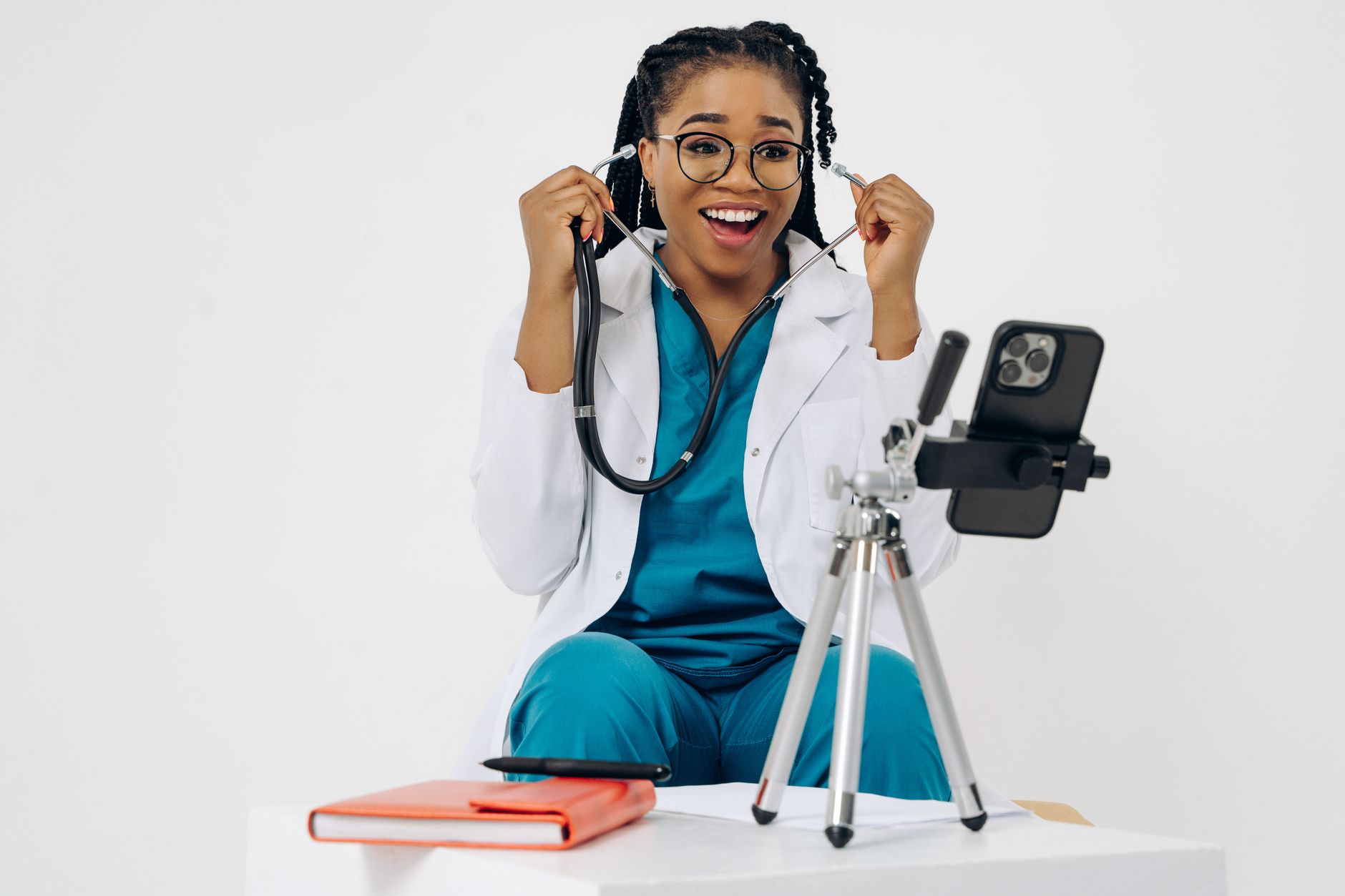 A cheerful female doctor in scrubs and white coat using a smartphone for a virtual medical consultation.
