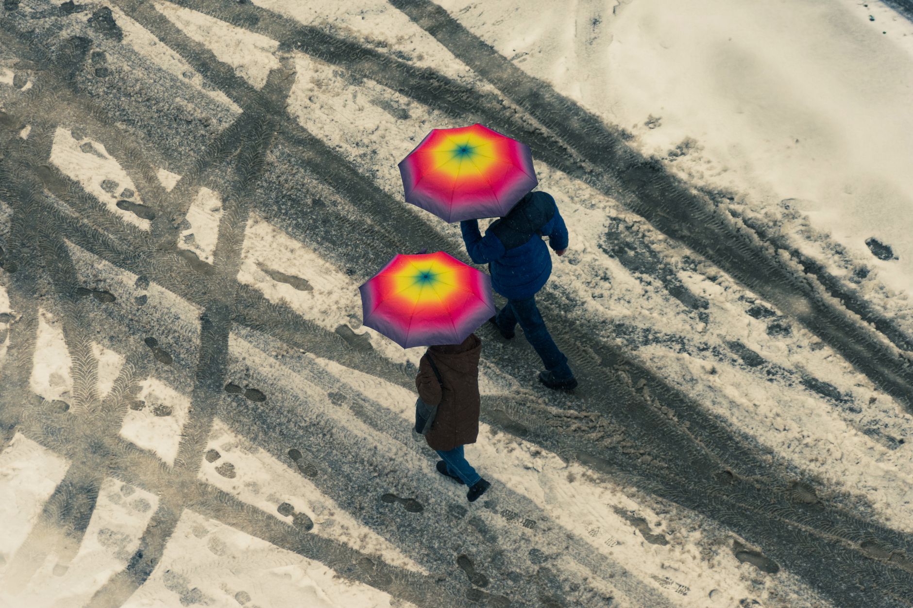 Two people walk on a snowy street with vibrant umbrellas, viewed from