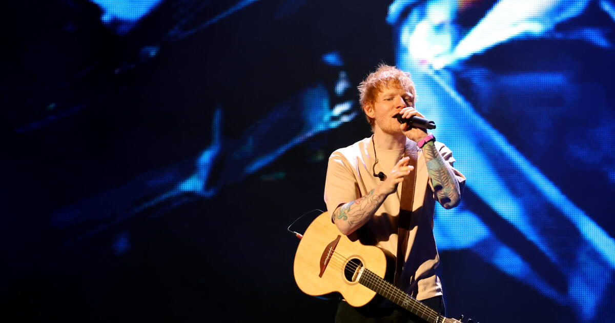 Ed Sheeran singing into a microphone. He wears a tan shirt, black pants and has a guitar slung over his body.