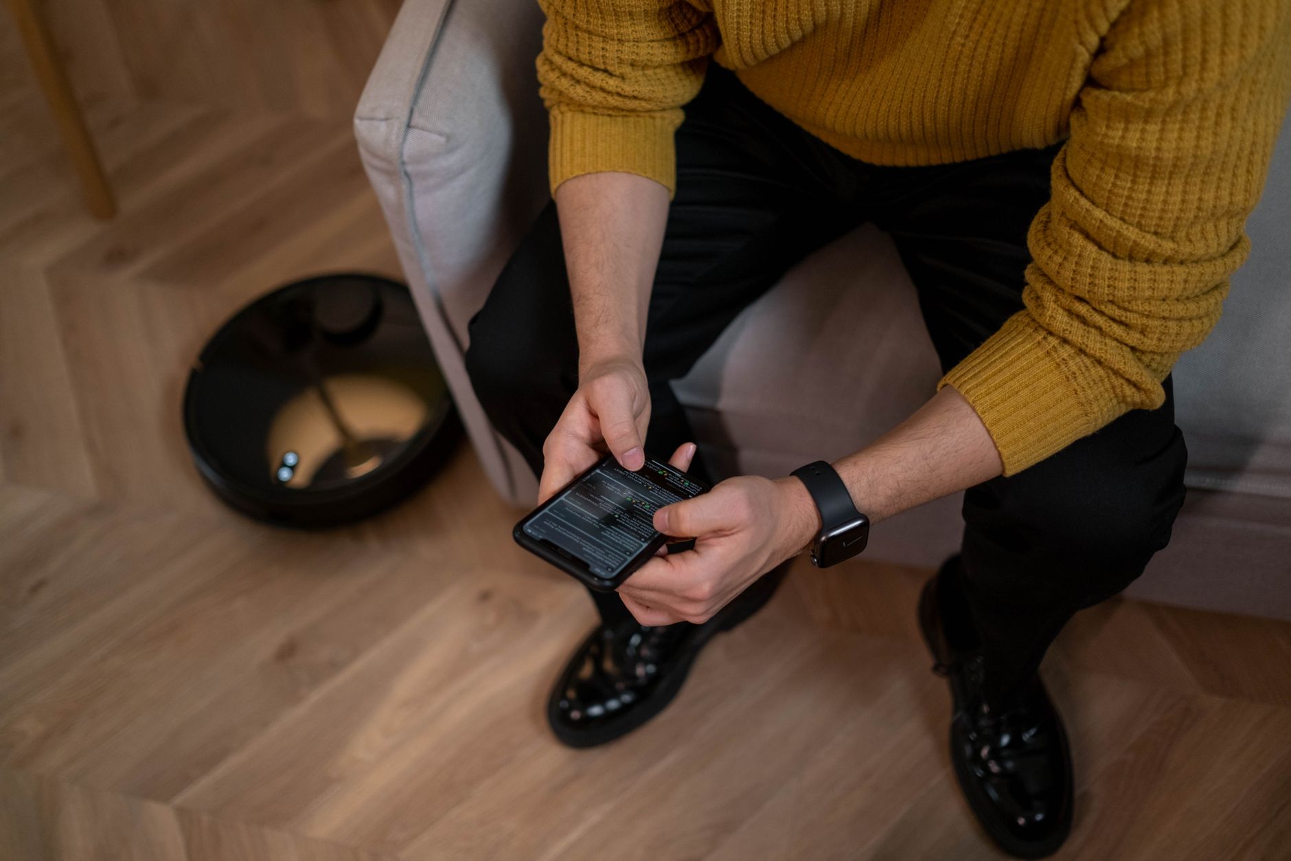 Man in yellow sweater using smartphone, robotic vacuum on wooden floor, modern
