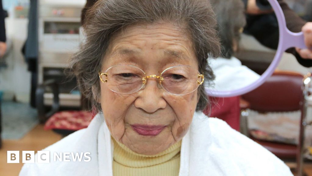 A 100-year-old Japanese woman wearing a butter yellow turtleneck, white jacket and rimless glasses at a hair salon with a hairdresser holding up a mirror behind her head