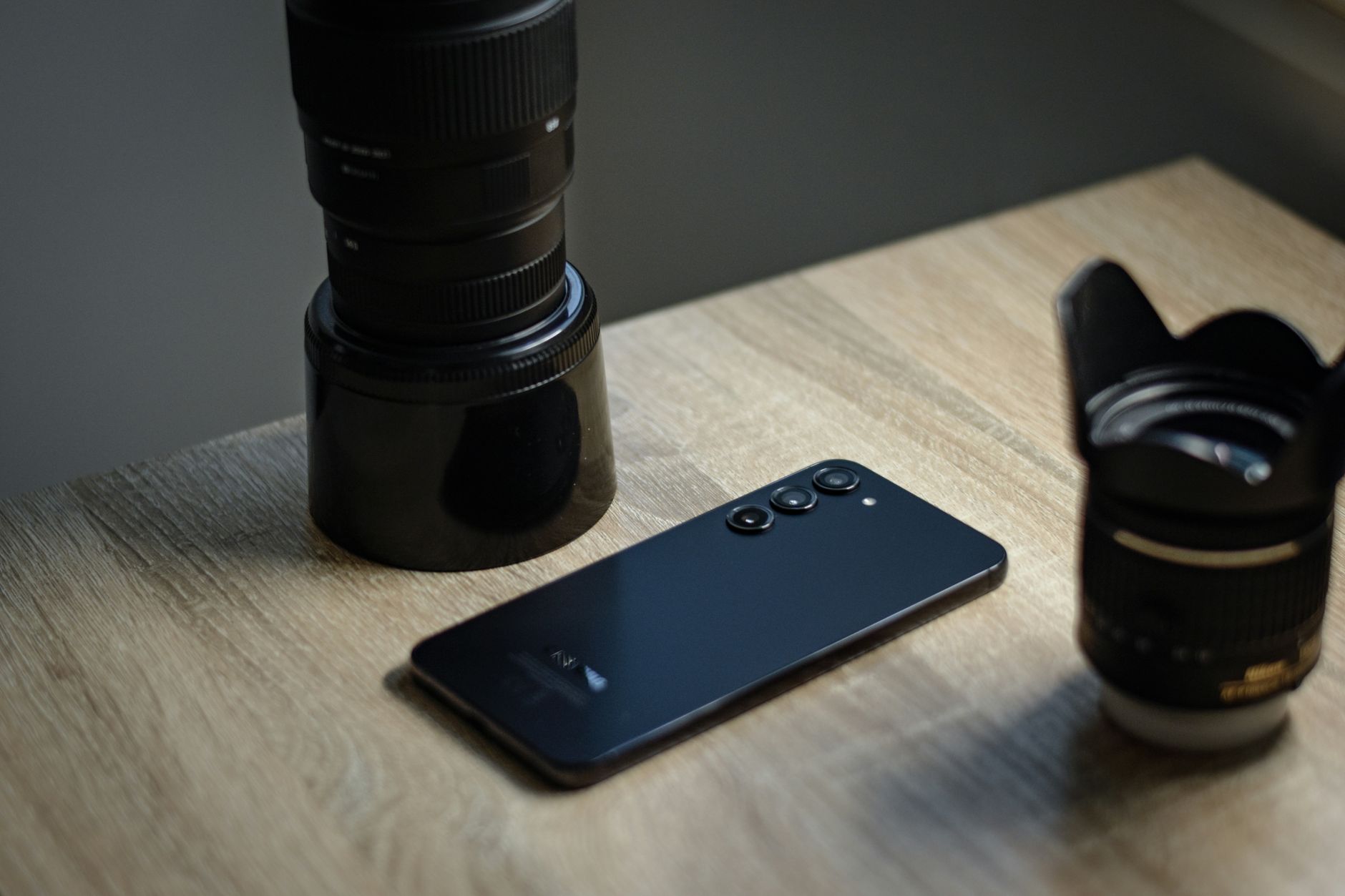 A sleek smartphone placed alongside camera lenses on a wooden desk, capturing