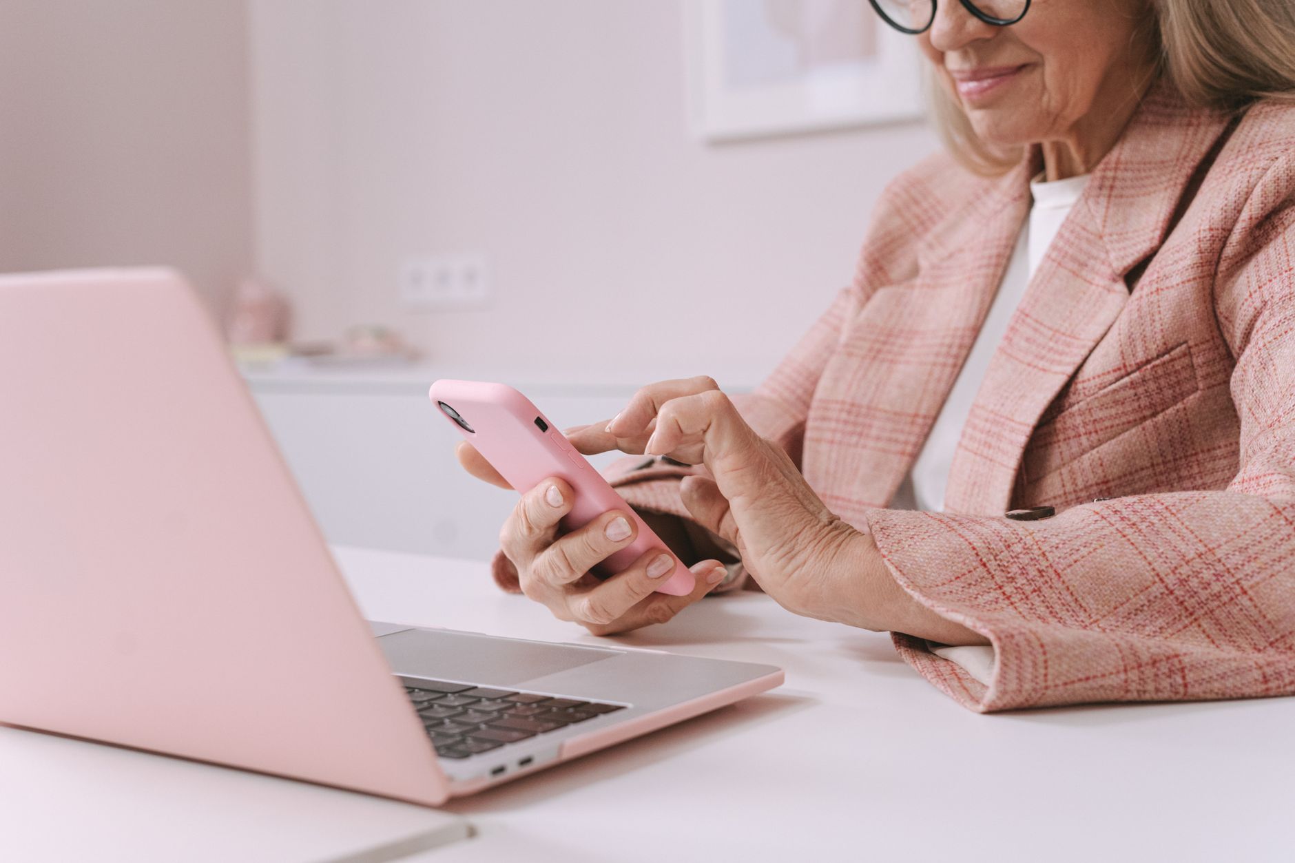 Elderly woman in pink plaid suit using a smartphone and laptop at