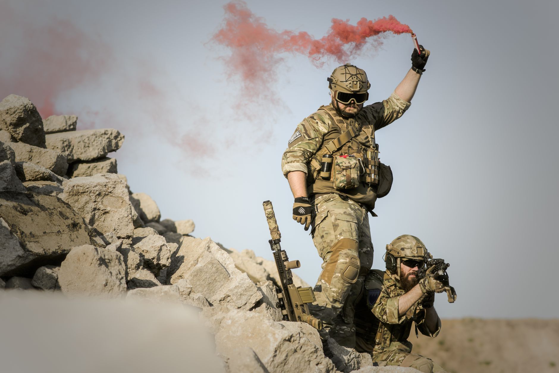 Two soldiers in camouflage, one signaling with red smoke in a desert
