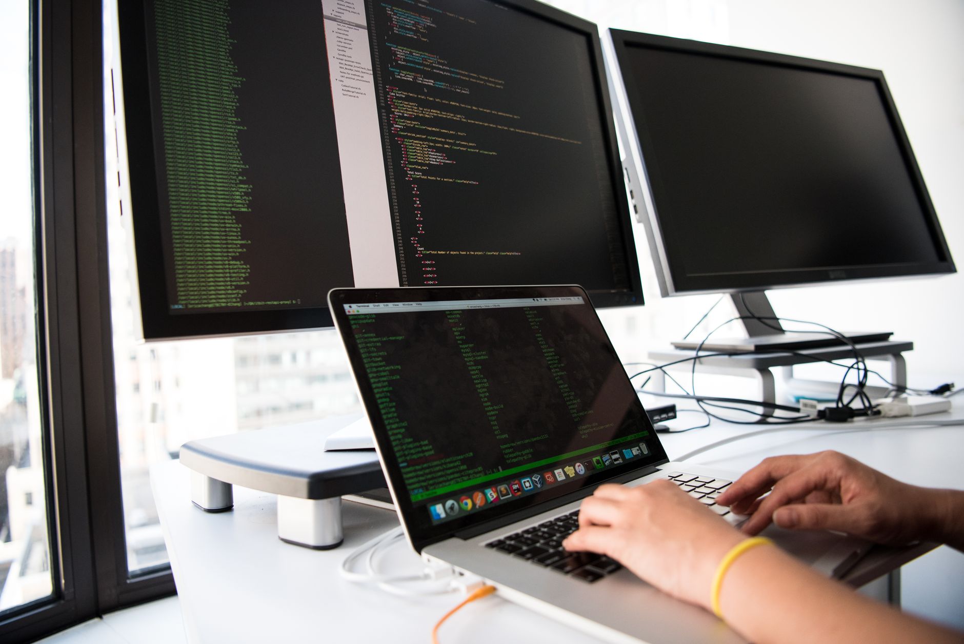 A developer writes code on a laptop in front of multiple monitors