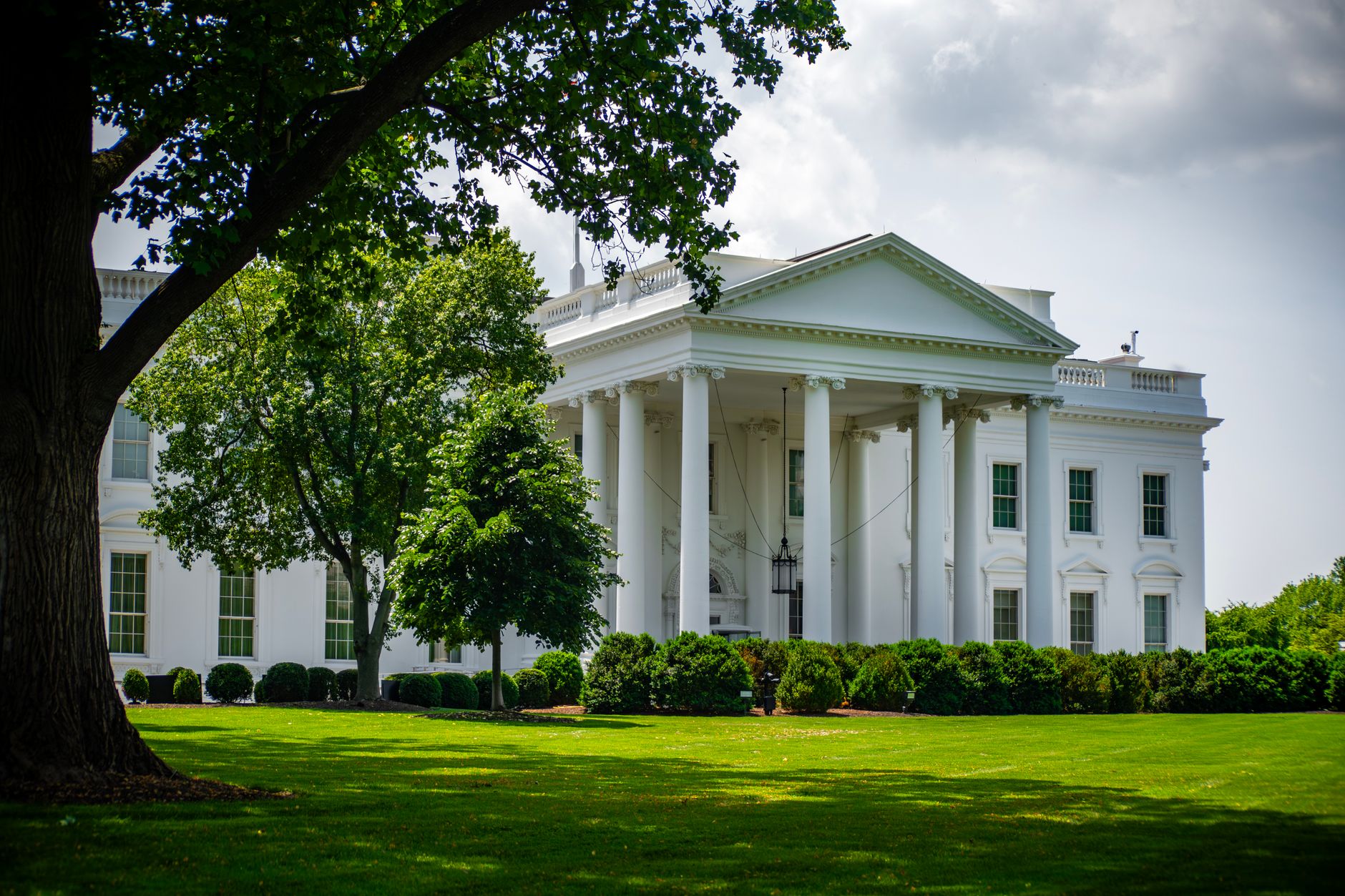 The White House framed by trees and greenery, in Washington, D.C., under a bright sky.