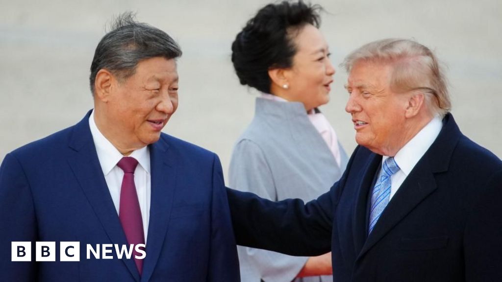 President Xi smiles and looks toward Trump, who has his hand on Xi's shoulder. Both are in suits and their haor is slightly tussled by wind. Xi's wife, in a light blue coat, looks away in the backgrou