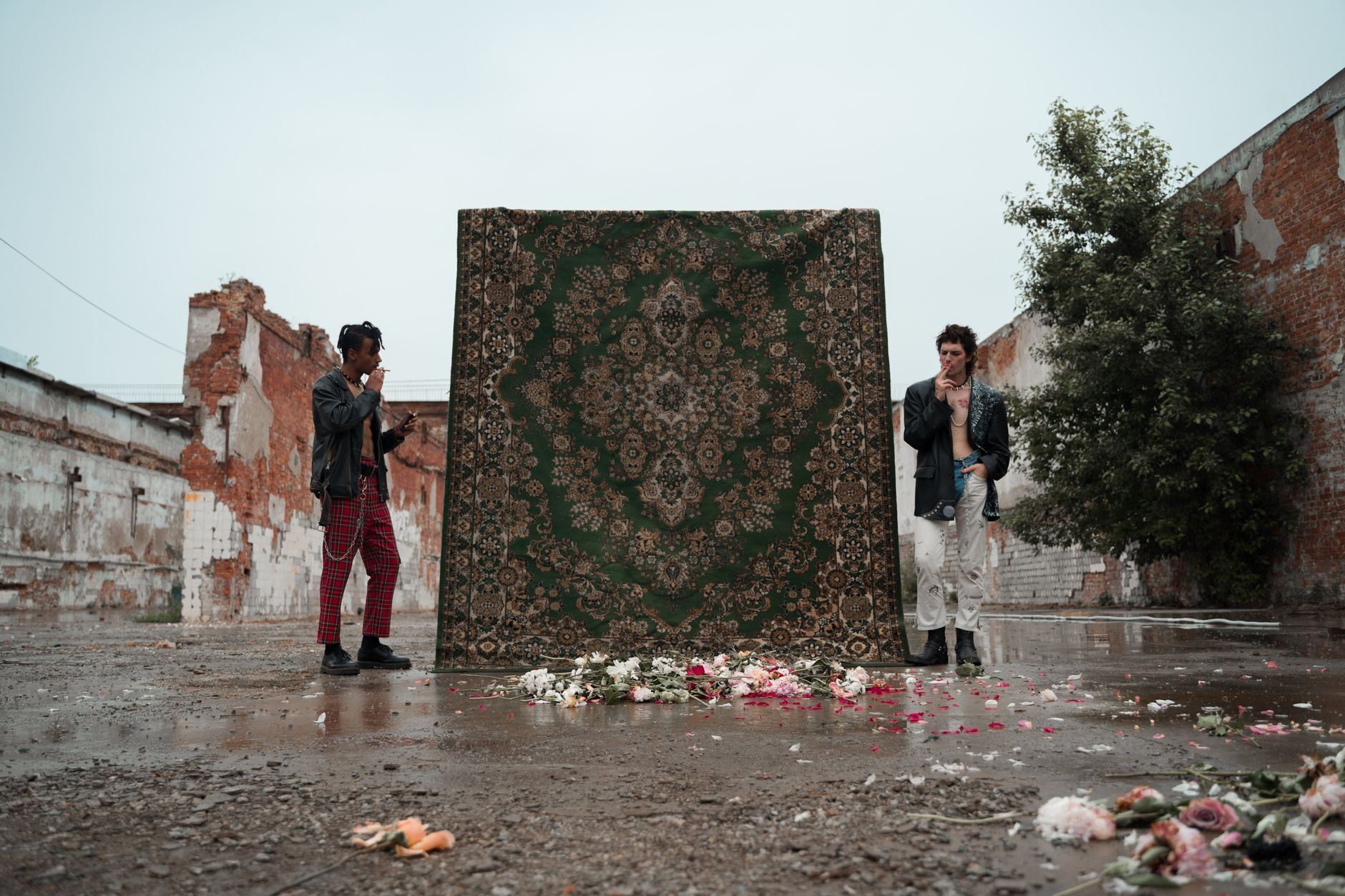 Two men in punk attire display a vintage carpet amid urban decay