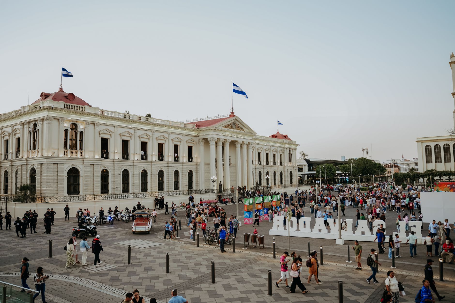 Crowded plaza scene in San Salvador with flags and historic buildings under