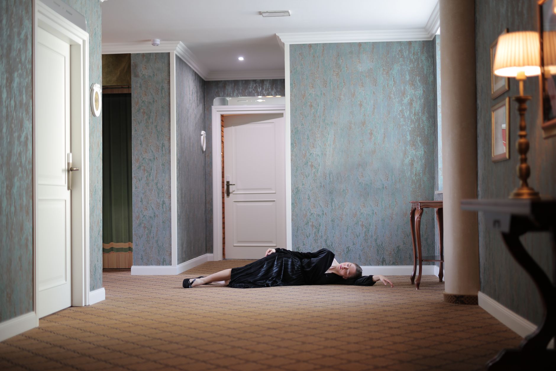 A woman in a black dress lies on the carpeted floor of an opulent hotel hallway.