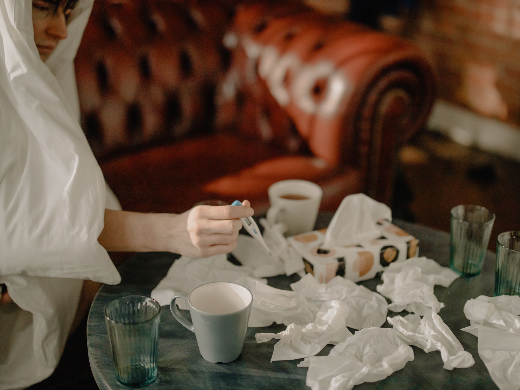 A man wrapped in a blanket checks his temperature, surrounded by tissues and cups.