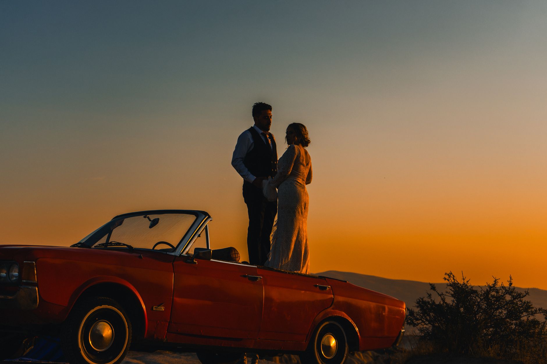A couple embraces on a vintage car at sunset, creating a romantic silhouette in Türkiye.