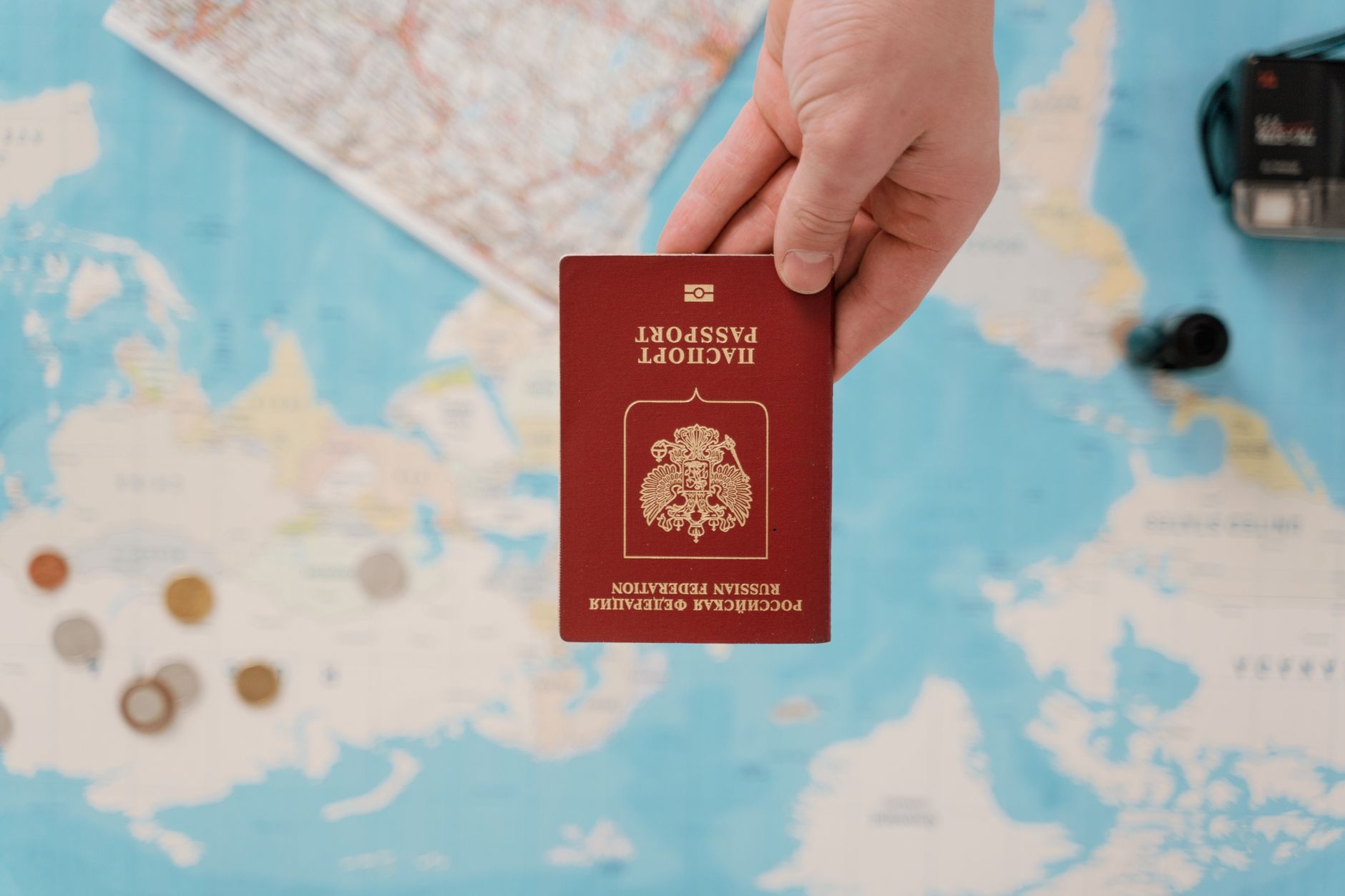 A hand holding a Russian passport above a travel-themed map, capturing world