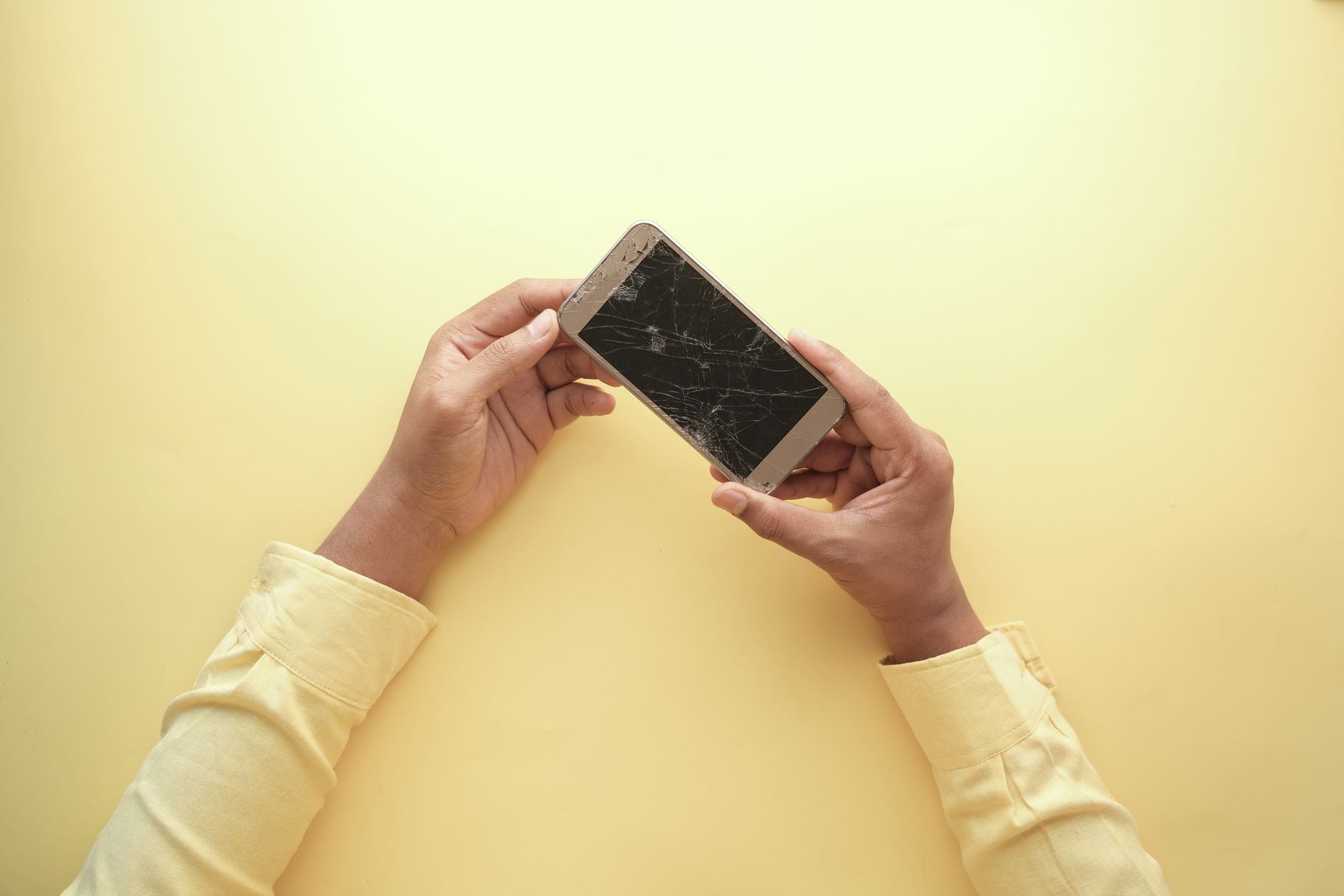Close-up of hands holding a cracked smartphone against a yellow backdrop, highlighting