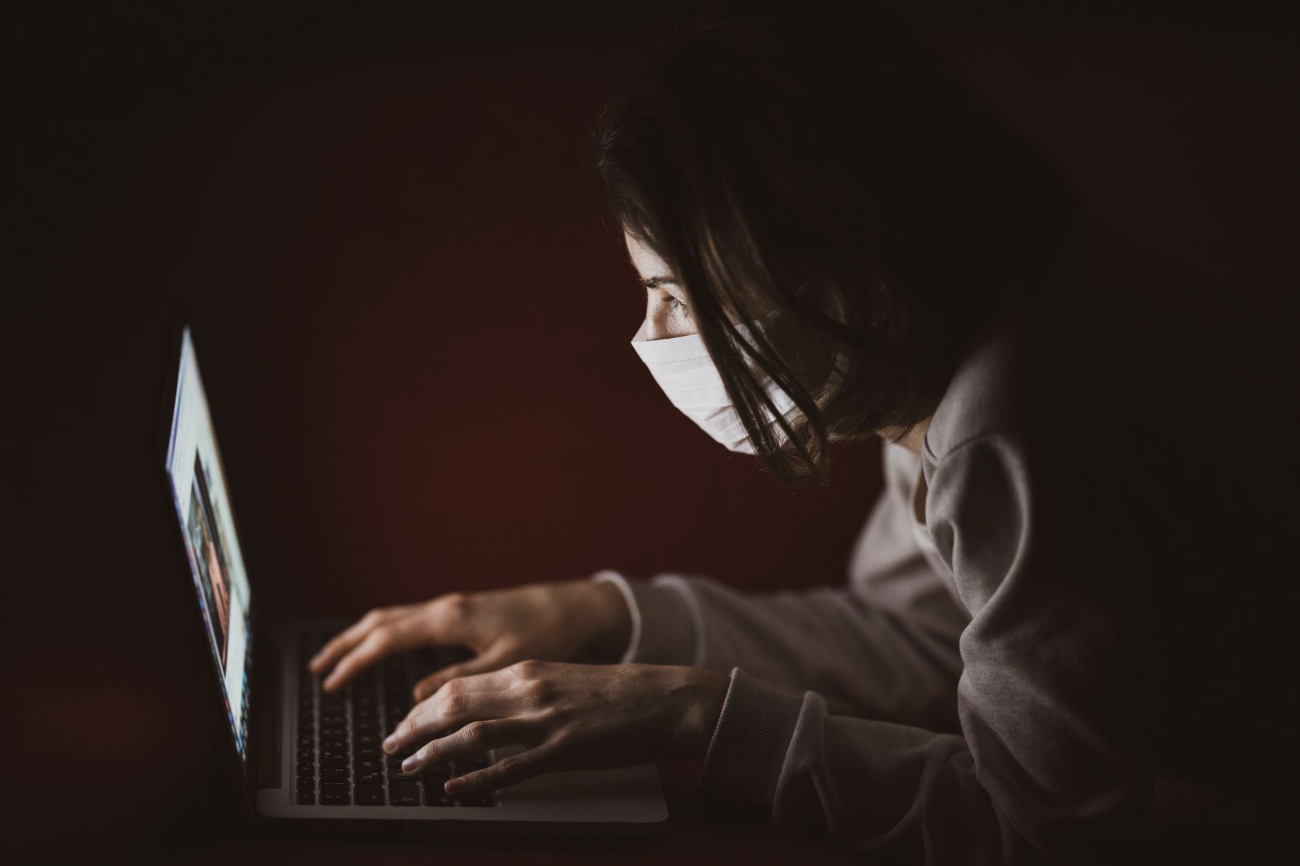 A woman wearing a face mask focuses on her laptop screen in dim lighting, highlighting remote work.