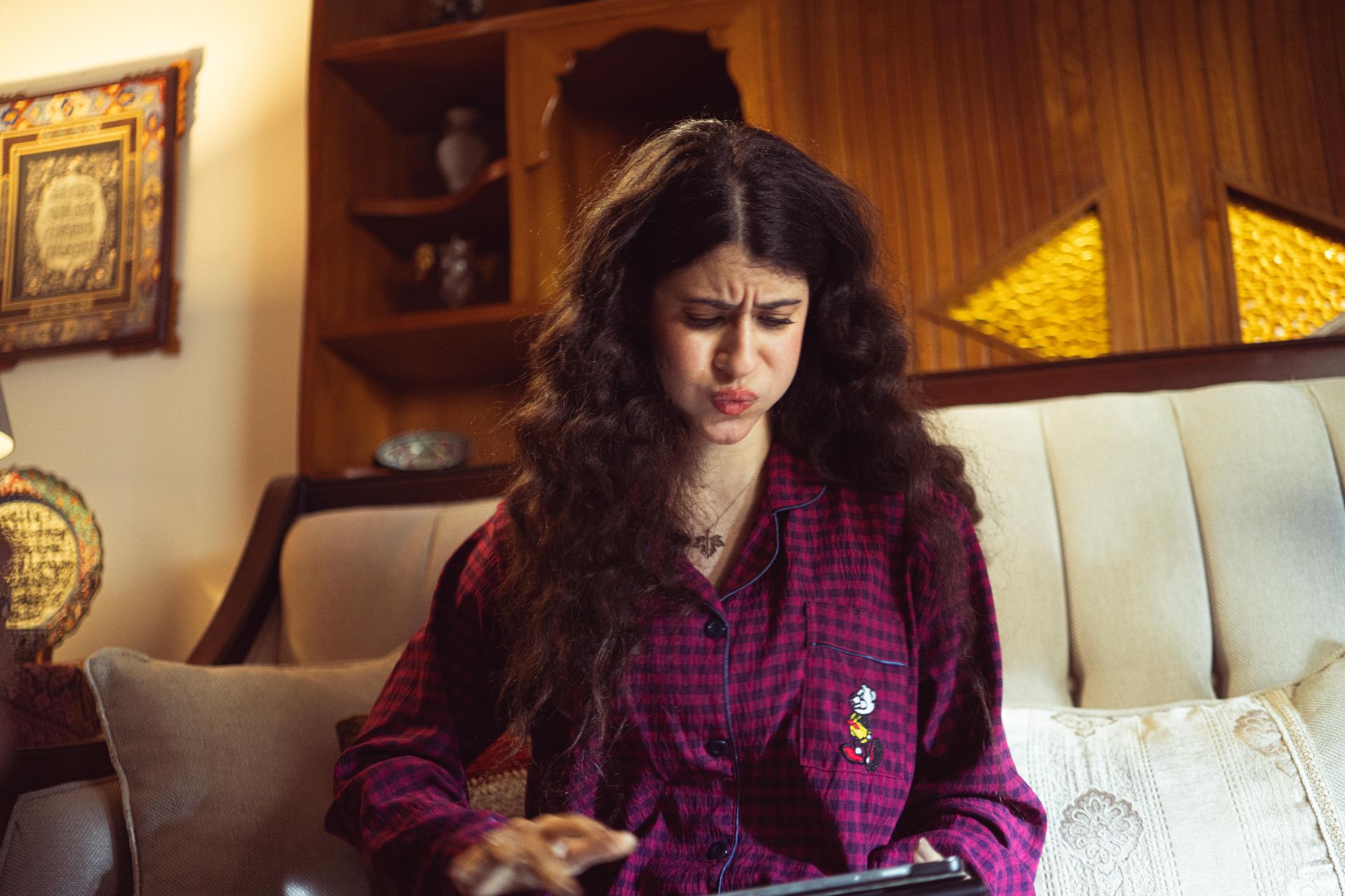 A woman sitting indoors looking frustrated with a tablet in hand, wearing
