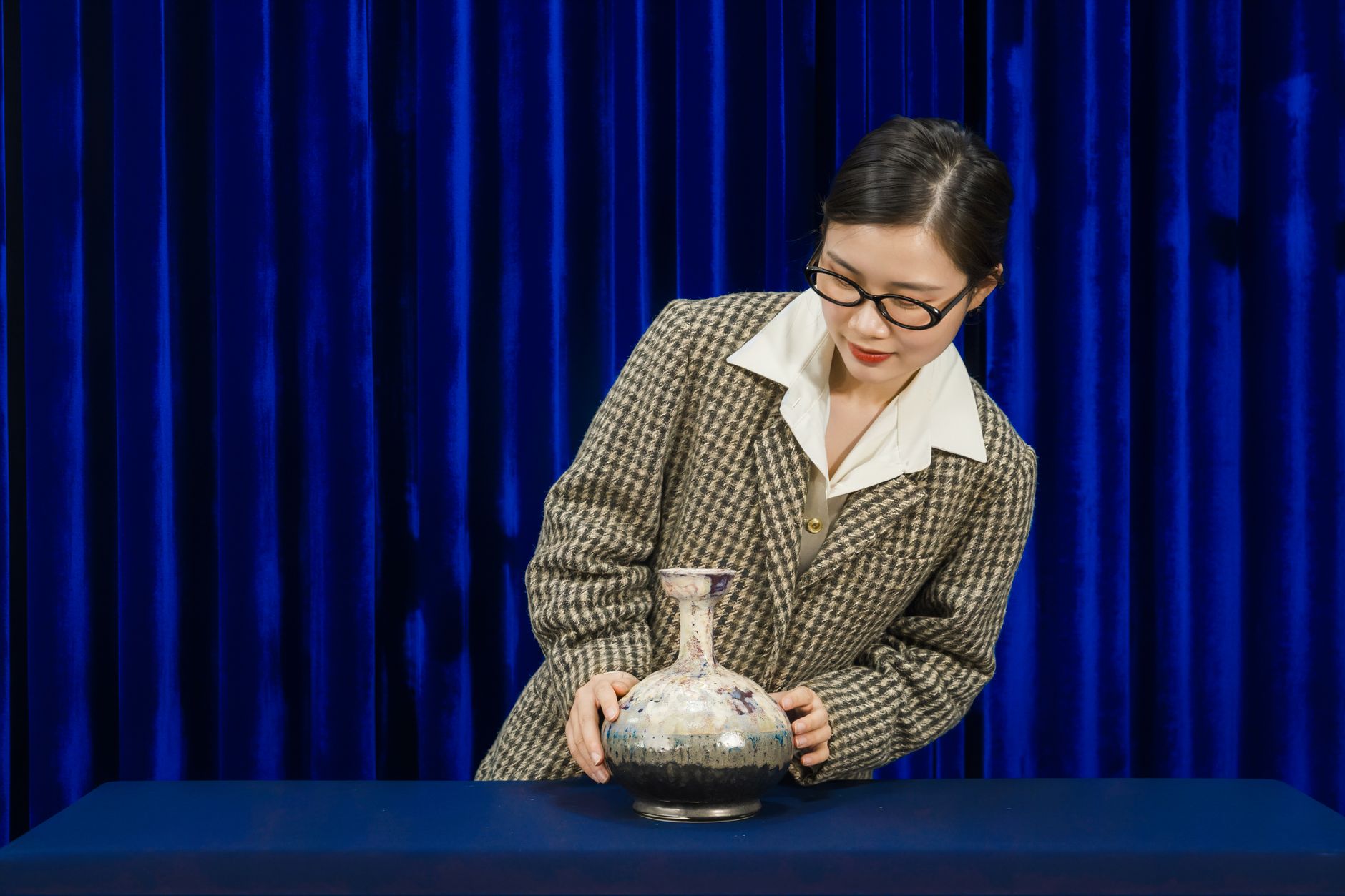 Woman examining a decorative vase in an elegant setting with a blue curtain backdrop.