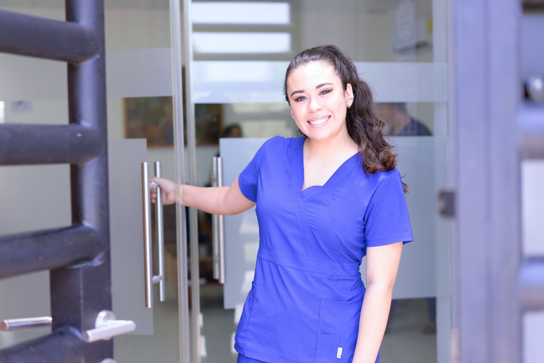 Smiling female nurse in blue scrubs opens a glass door in a