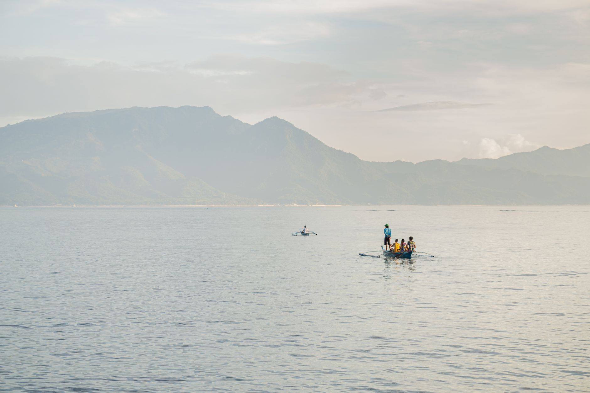 People enjoying a calm canoe ride on a vast ocean with mountains under a bright sky.