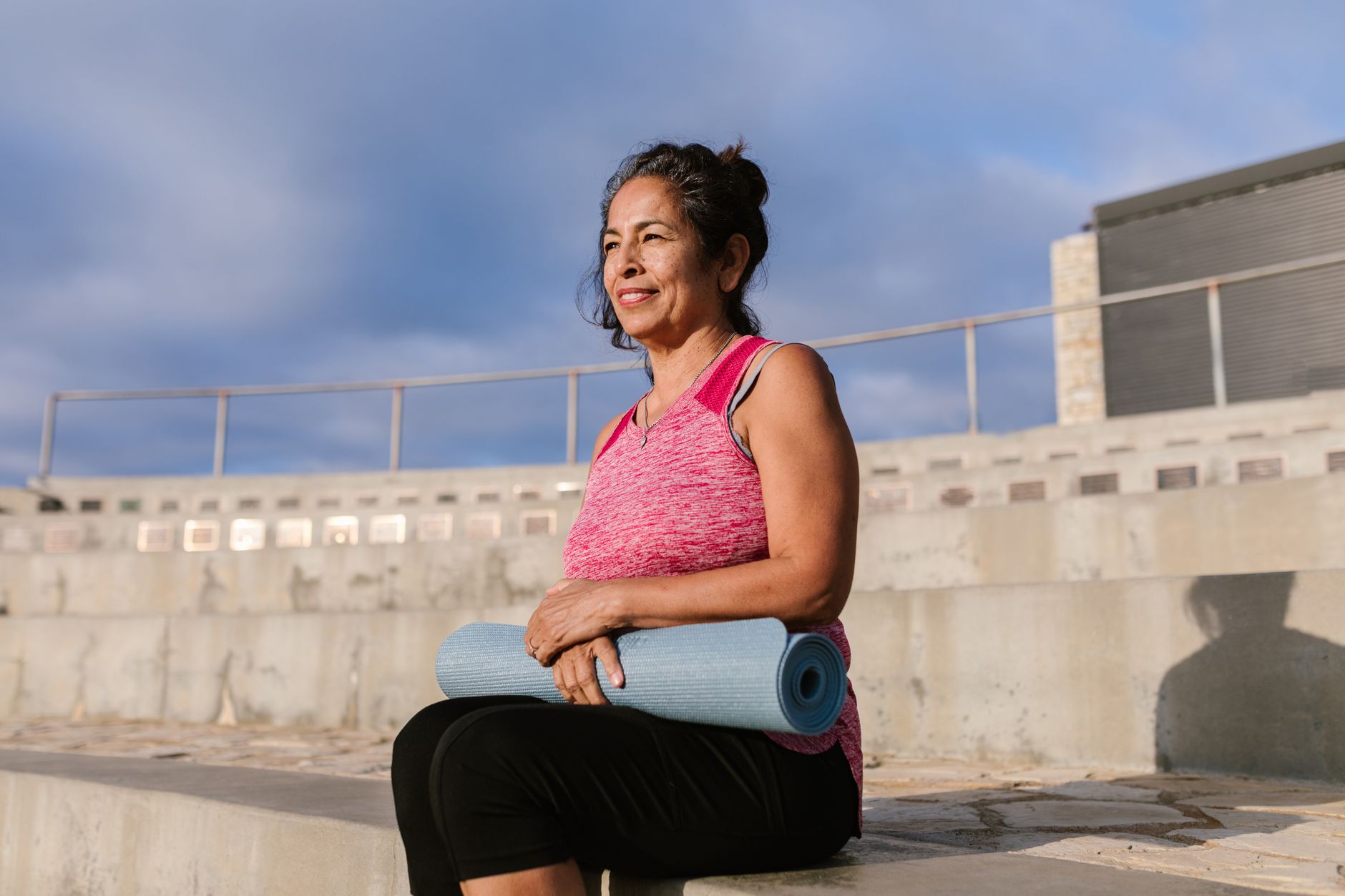 Middle-aged woman holding a yoga mat while enjoying the outdoors. Promotes a