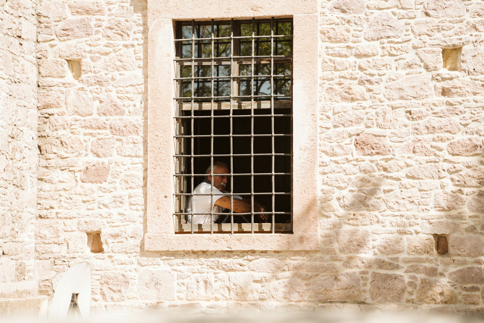 An elderly man is seen sitting inside a building, framed by a barred window of a stone wall.