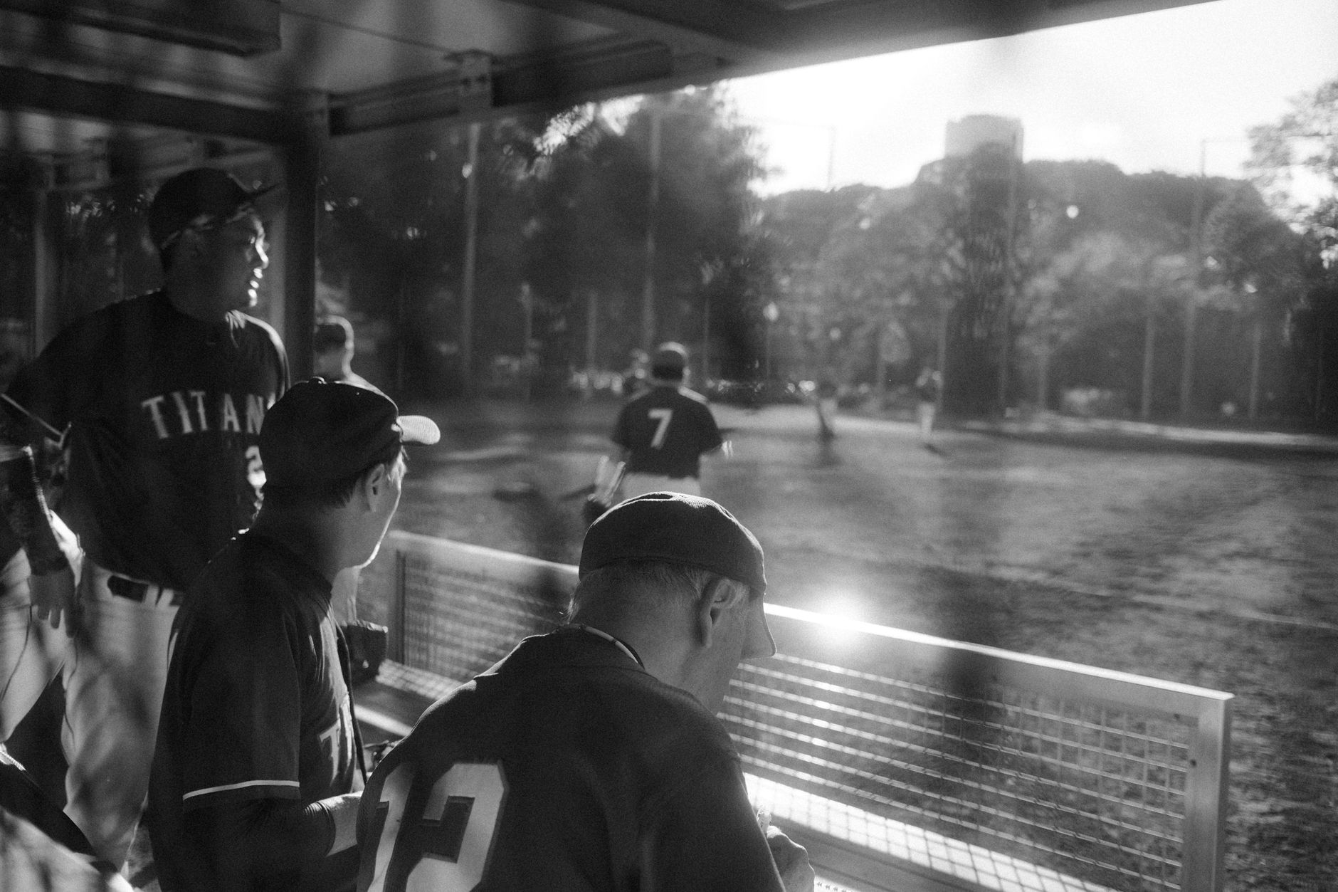 Baseball players in a dugout during a game in a sunlit Tokyo park.