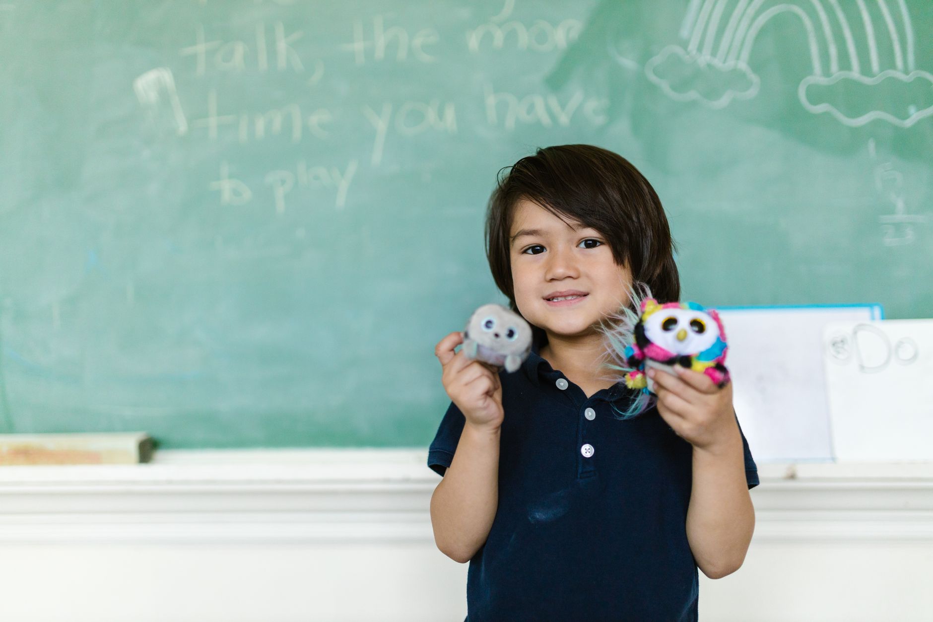 Smiling child holds plush toys in a classroom setting, with chalkboard background.