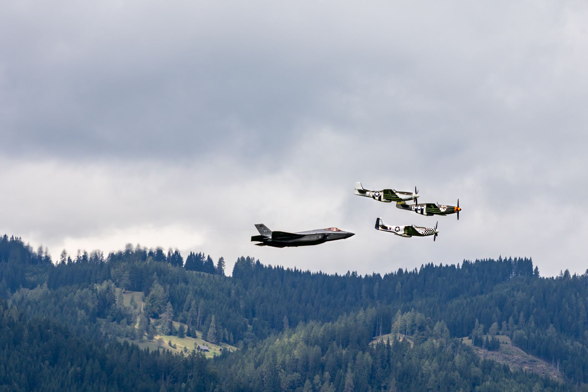 Three military aircraft in formation fly over a dense forest under a