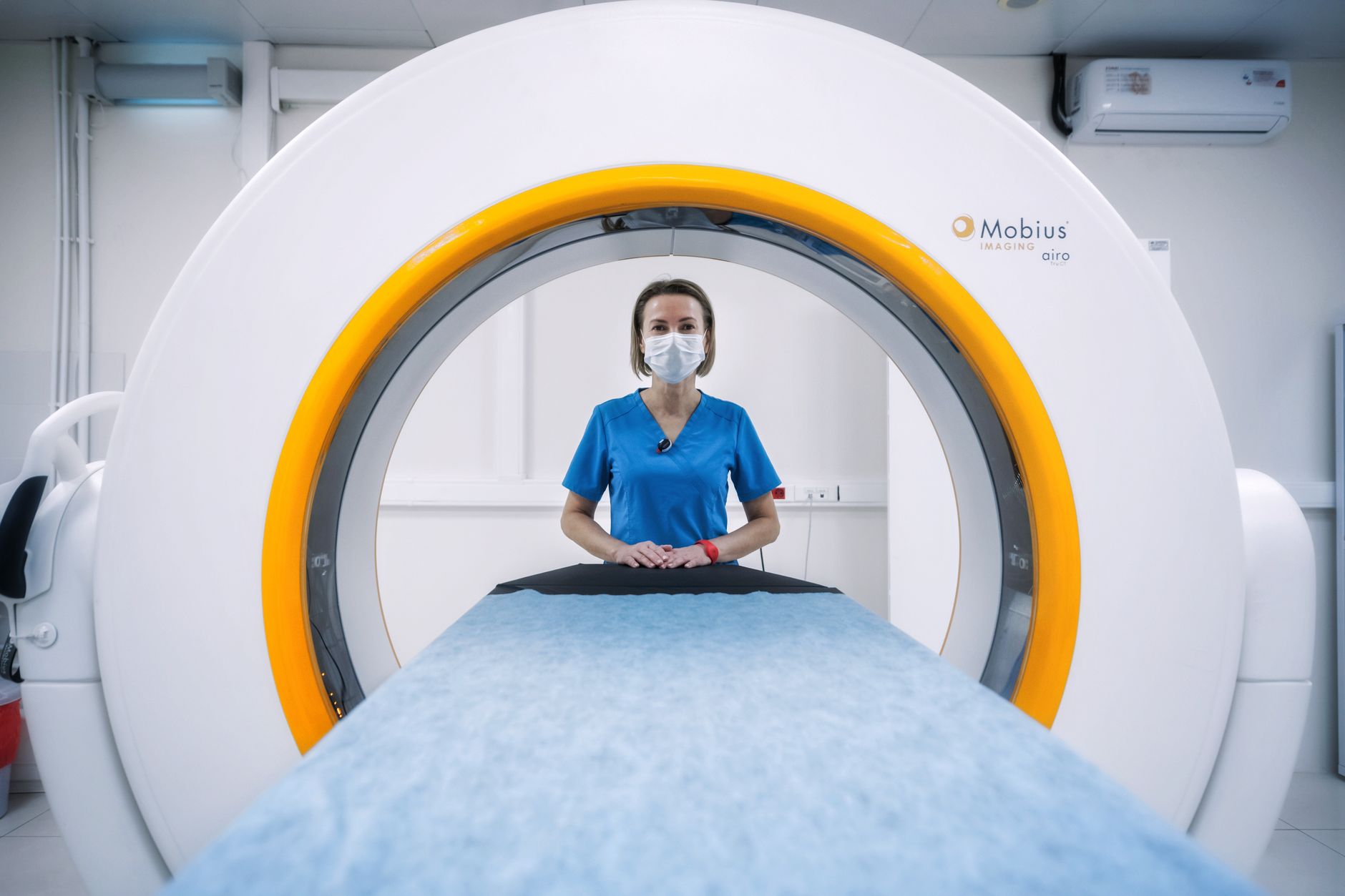 Nurse in blue scrubs and mask stands by an MRI machine, ready for a scan.