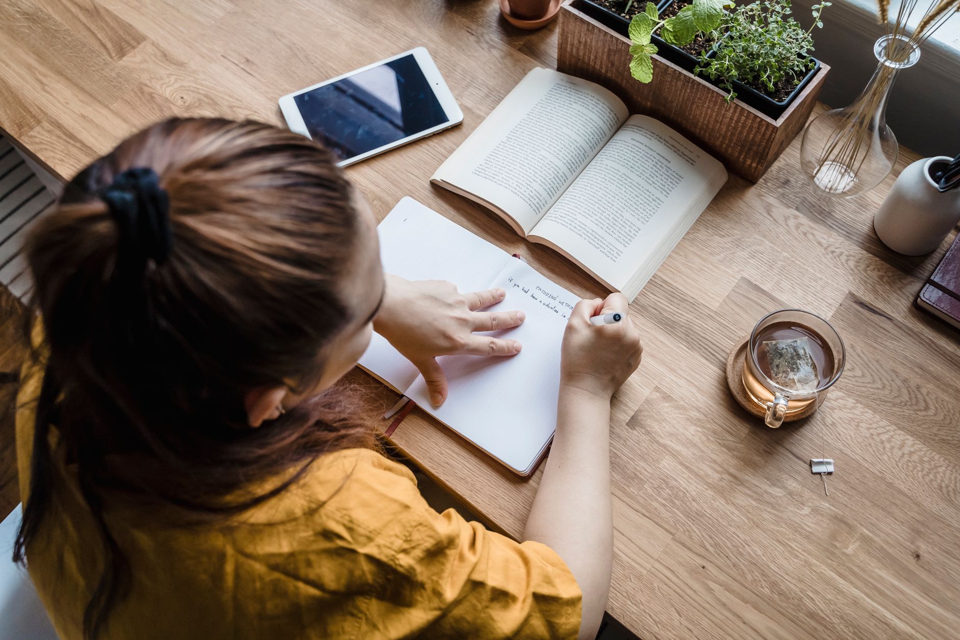 A woman writing in a notebook at a wooden desk with books,
