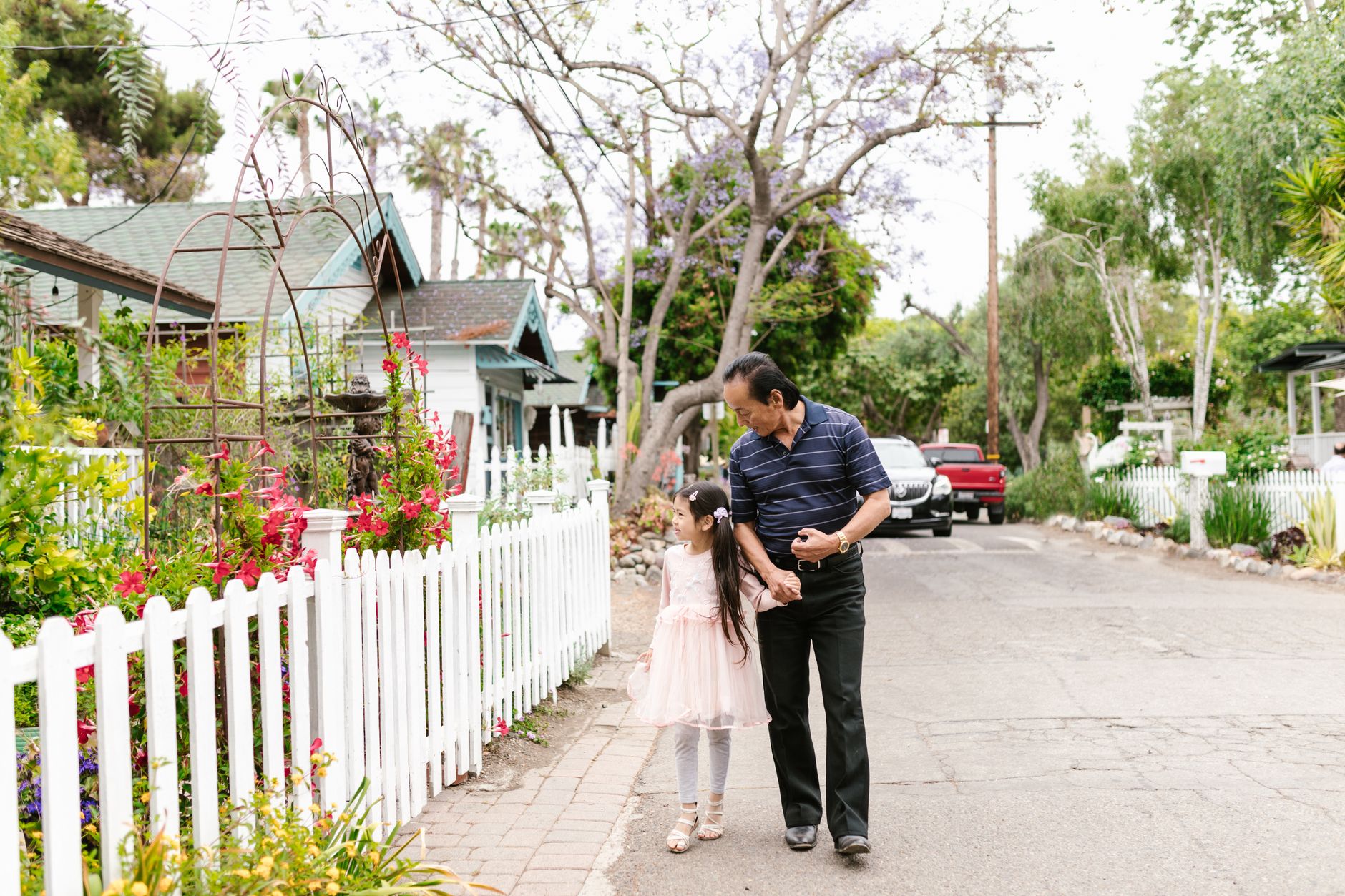 A senior man and young girl walking hand in hand on a suburban street lined with white fences.