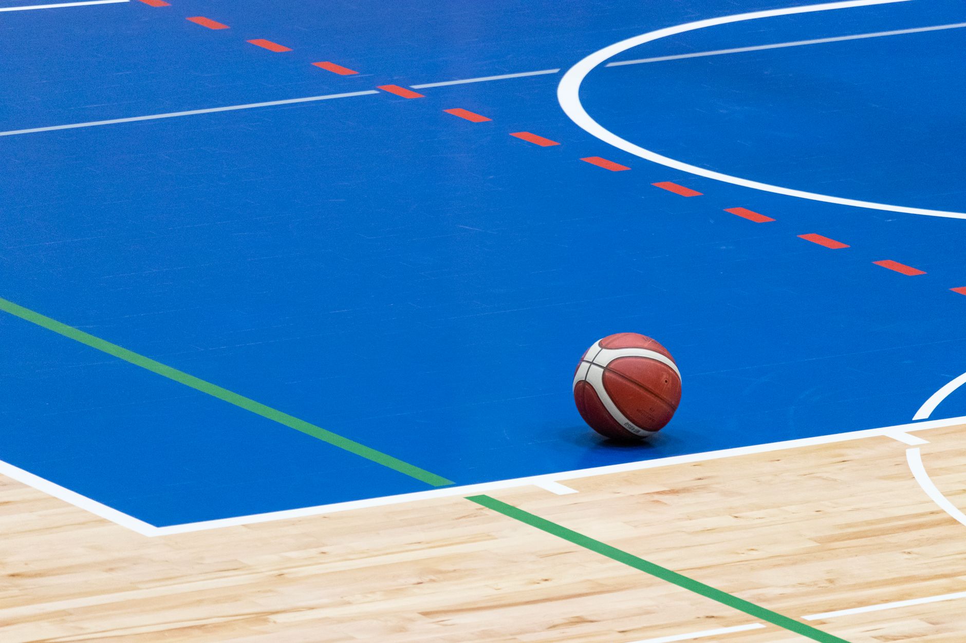 A basketball rests on a vibrant blue indoor court with wood flooring and colorful lines.